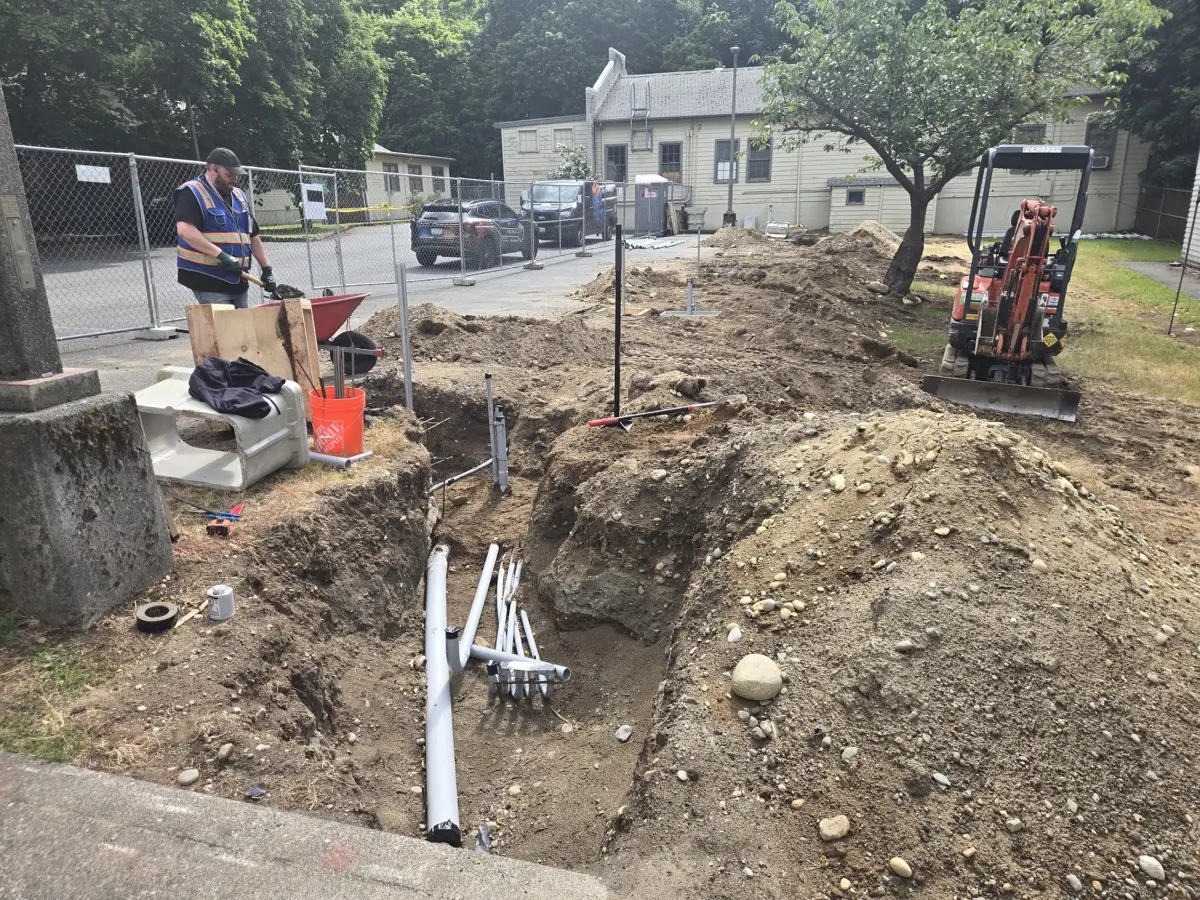 Crew hand-digging a conduit trench near the building foundation